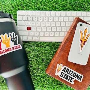 May include: Three Arizona State University stickers. One sticker features the school's mascot, the Sun Devil, with a spear. Another sticker features the school's name and three hands making the peace sign. The third sticker features the school's name and a spear.