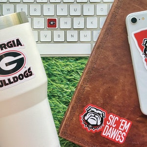 May include: A white tumbler with a black and green Georgia Bulldogs logo sticker. A brown leather journal with a red and white bulldog sticker that says "Sic 'Em Dawgs" and a white iPhone with a red and white Georgia bulldog sticker that says "Est. 1785".