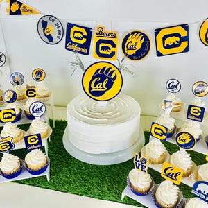 May include: A white frosted cake with a large "Cal" topper, surrounded by cupcakes and decorations. The cupcakes and decorations feature the "Cal" logo and bear paw prints in blue and gold. A banner with "Go Bears" is visible.