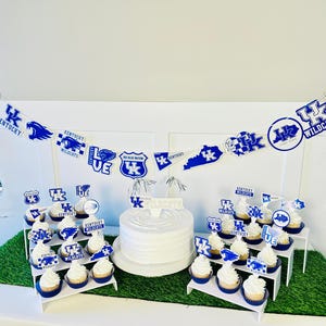 Może przedstawiać: Biały tort z niebiesko-białym lukrem i niebiesko-białym banerem z napisem "Kentucky Wildcats" i "Go Blue Wildcats". Na stojaku warstwowym znajdują się babeczki z niebiesko-białym lukrem i niebiesko-białymi ozdobami.