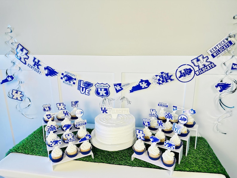 Może przedstawiać: Biały tort z niebiesko-białym lukrem i niebiesko-białym banerem z napisem "Kentucky Wildcats" i "Go Blue Wildcats". Na stojaku warstwowym znajdują się babeczki z niebiesko-białym lukrem i niebiesko-białymi ozdobami.