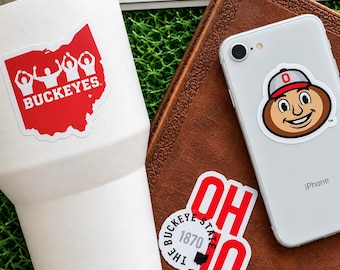 May include: A white stainless steel tumbler with a red Ohio-shaped sticker featuring silhouettes of people and the text BUCKEYES. A silver smartphone with a Brutus Buckeye sticker and a red OH-IO sticker with 1870 text on a brown leather notebook.