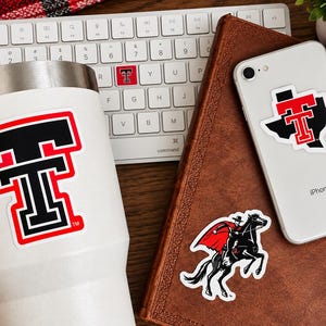May include: A white tumbler, a silver smartphone, and a brown notebook featuring Texas Tech University stickers. The stickers display the red and black double T logo, the state of Texas with the double T, and a masked rider on a black horse.