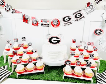 May include: A white cake decorated with a red and black Georgia Bulldogs logo, surrounded by cupcakes with red and black Georgia Bulldogs themed toppers. The cupcakes are arranged on three tiers of white cupcake stands. The background features a white wall with a red and black Georgia Bulldogs themed banner.