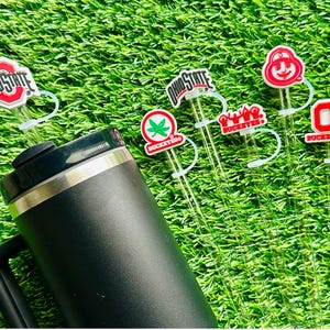 May include: Six different straw toppers with various Ohio State University logos and designs. The toppers are red, white, and black. The straws are clear and are inserted into a black travel mug with a handle.