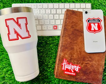 May include: White tumbler with a red "N" logo, a brown leather journal with a red "Huskers" logo and a white phone case with a red "Nebraska N" logo.
