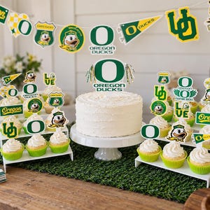 May include: A white frosted cake on a pedestal stand surrounded by cupcakes in green liners, all decorated with Oregon Ducks themed paper toppers and banners featuring the team logo, mascot, and text in green, yellow, and white colors.