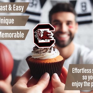 May include: A cupcake with white frosting and chocolate sprinkles topped with a red and black University of South Carolina Gamecocks logo cupcake topper. The cupcake is being held by a person's hand. The background is blurred and shows a person wearing a white shirt with a black and white striped pattern.