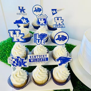 Może przedstawiać: Tr&oacute;jpoziomowa podstawka na babeczki z niebieskimi i białymi babeczkami w temacie Kentucky Wildcats. Babeczki są ozdobione lukrem i dodatkami przedstawiającymi logo drużyny, maskotkę i inne powiązane obrazy. Podstawka jest otoczona zieloną trawą.