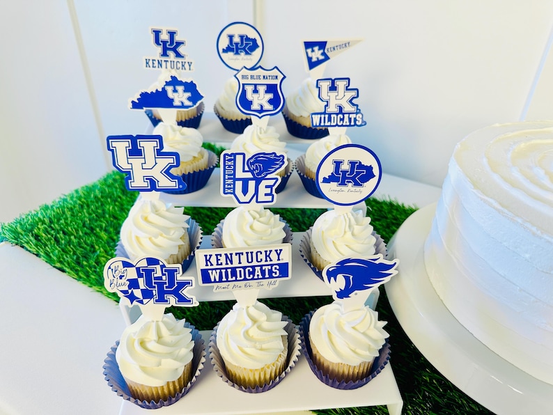 Może przedstawiać: Tr&oacute;jpoziomowa podstawka na babeczki z niebieskimi i białymi babeczkami w temacie Kentucky Wildcats. Babeczki są ozdobione lukrem i dodatkami przedstawiającymi logo drużyny, maskotkę i inne powiązane obrazy. Podstawka jest otoczona zieloną trawą.