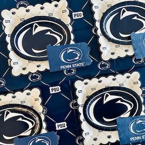 May include: Penn State party supplies, including plates and napkins, are arranged on a blue patterned surface. The plates have a white scalloped edge and feature the Penn State logo in navy blue and white. Napkins in the shape of Pennsylvania also display the logo.