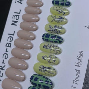 Puede incluir: Colección de uñas artificiales con varios diseños. Algunas son de color nude, otras amarillo claro con dibujos lineales negros. Otras presentan patrones de cuadros verdes y azules. El texto "NÄL ART" es visible.