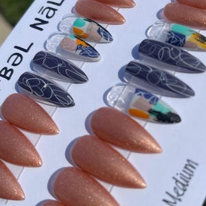 Puede incluir: Una colección de uñas postizas en forma de estilete. Algunas uñas son de color oro rosa brillante, mientras que otras presentan diseños abstractos en azul, blanco, naranja y turquesa. El texto "Böl Näl Är" es visible en la esquina superior izquierda y "Stiletto Medium" en la inferior derecha.