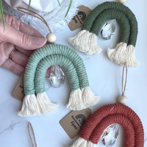 May include: Three macrame rainbow ornaments in sage green, olive green, and terracotta. Each arch features a central clear crystal teardrop prism, a natural wooden bead at the top, and a jute hanging cord. Each includes a brown paper tag.
