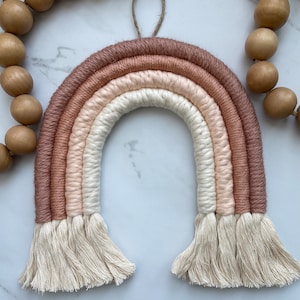 May include: A macrame rainbow wall hanging with a neutral color palette of blush pink, peach, and cream. The rainbow is made of thick, twisted yarn and has a fringe at the bottom.