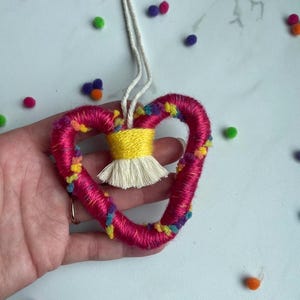 May include: A handmade heart-shaped ornament with a bright pink yarn exterior, accented with colorful pom-poms. The center features a yellow wrapped section with a white tassel. A white string is attached for hanging. Scattered colorful pom-poms surround the ornament.