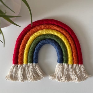 May include: A handmade rainbow wall hanging. The rainbow features red, orange, yellow, green, and blue arches, with a cream-colored fringe at the base. The craft is made of yarn and is displayed on a white surface.