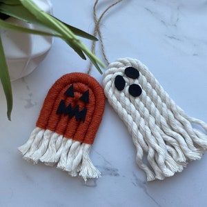 May include: Two macrame Halloween decorations: a pumpkin and a ghost. The pumpkin is orange with a black jack-o'-lantern face. The ghost is white with three black felt eyes. Both have a fringe at the bottom and a twine loop for hanging.