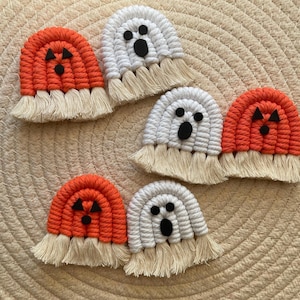 May include: Macrame Halloween decorations featuring orange pumpkins and white ghosts with black facial features. Each piece has a fringed bottom and is set against a woven background.