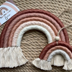 May include: Two macrame rainbows in shades of brown, pink, and white with fringe. The larger rainbow is hanging from a string with a wooden bead. The smaller rainbow is on the table. The card reads "Rainbows for Lynn".