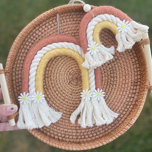 May include: A handmade macrame rainbow wall hanging in shades of peach, yellow, and cream, adorned with white daisy accents. The rainbow is suspended on a woven rattan base with wooden handles, creating a charming, bohemian-style decor piece.