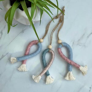 May include: Three handmade macrame ornaments in the shape of a ribbon. The ornaments are made with blue, pink, and teal yarn and have wooden beads at the top.