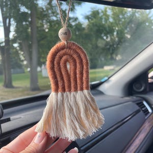 May include: A handmade car air freshener with a woven rainbow design in brown and cream. The rainbow hangs from a wooden bead with the word "LOVE" engraved, suspended by twine. The air freshener is designed for a car.
