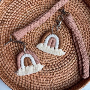 May include: Two macrame keychains featuring braided wrist straps in a dusty rose color. Each keychain includes a small rainbow charm made of white, dusty rose, and brown cord with a fringed bottom, attached by a silver metal swivel lobster clasp.