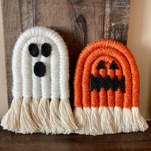 May include: Two macrame Halloween decorations: a white ghost with black eyes and mouth, and an orange pumpkin with a black face. Both have a fringe at the bottom. The decorations are made of yarn and are set against a wooden background.