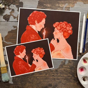 Puede incluir: Dos impresiones artísticas que presentan una ilustración romántica de una pareja con vestimenta de época, iluminada por la luz de las velas. Las impresiones son en tonos rojos y melocotón sobre un fondo oscuro. La obra de arte se exhibe sobre una superficie de madera con materiales de arte.