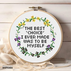 Puede incluir: Un bastidor de bordado circular de madera que contiene tela blanca con un diseño de punto de cruz. El texto dice THE BEST CHOICE I EVER MADE WAS TO BE MYSELF en negro, rodeado por una corona de pequeñas flores amarillas, moradas y verdes.