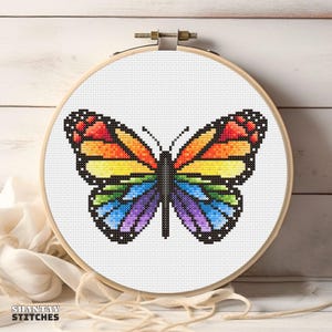 May include: A circular wooden embroidery hoop containing a cross-stitch pattern of a butterfly with rainbow-coloured wings. The butterfly features segments of red, orange, yellow, green, blue, and purple, outlined in black against a white fabric.