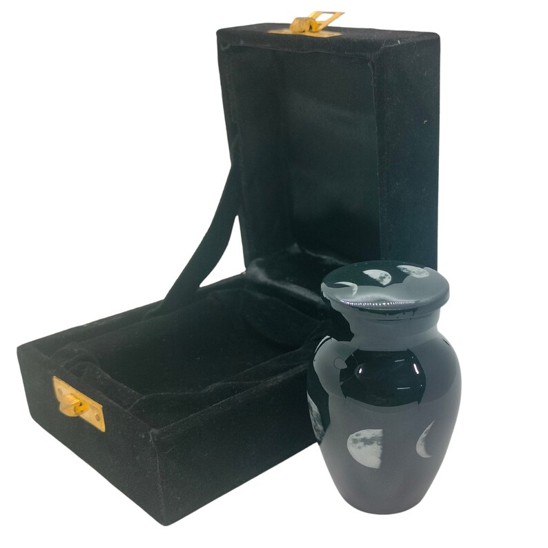 May include: Black cremation urn with a moon phases design. The urn is made of metal and comes in a black velvet box.