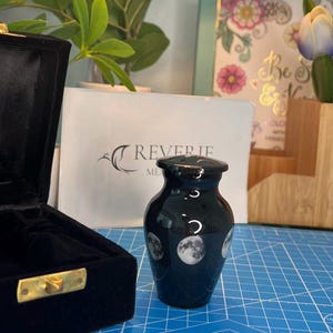 May include: Black cremation urn with a moon and stars design. The urn is made of ceramic and has a glossy finish. It is sitting on a blue surface.