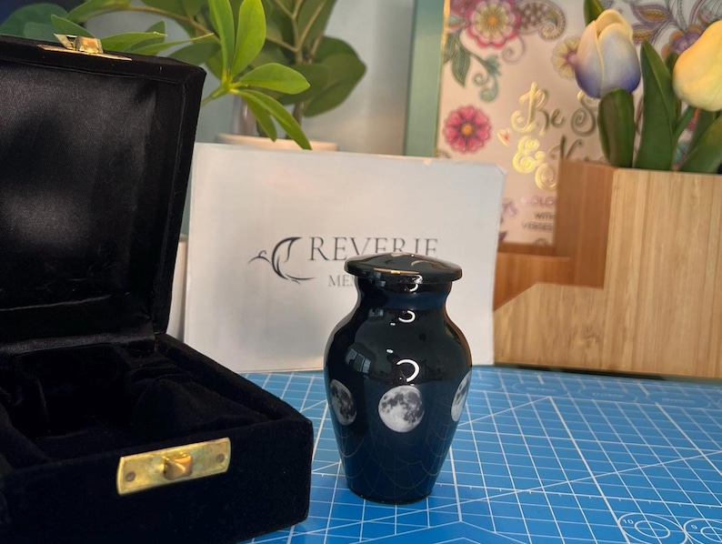 May include: Black cremation urn with a moon and stars design. The urn is made of ceramic and has a glossy finish. It is sitting on a blue surface.