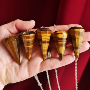 May include: Five tiger's eye crystal pendulums with silver chains. Each pendulum is a faceted, cone-shaped gemstone with bands of brown and gold. The pendulums are held in a hand against a dark red background.