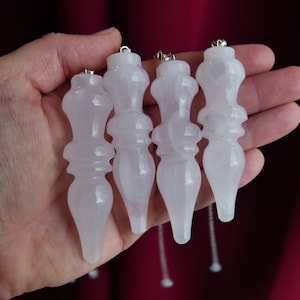 May include: Four pale pink, translucent, carved stone pendulums with a smooth, polished finish. Each piece features a tiered, spindle-like shape with a pointed base and a small silver-toned metal loop attached to the top for hanging or attachment.