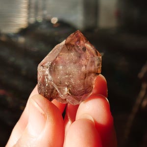 May include: A faceted, natural crystal with a pointed tip. The crystal has a translucent appearance with shades of brown and purple. The crystal is held in a hand, with the background out of focus.