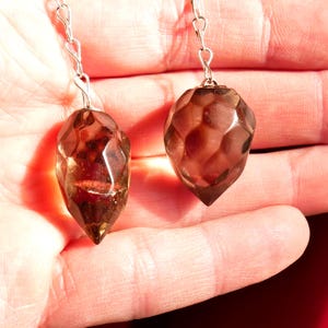 May include: Two faceted, teardrop-shaped smoky quartz pendants suspended from silver-toned metal chains. Each gemstone features a polished, multi-faceted surface with a translucent, dark brown hue that catches the light, creating depth and internal reflections.