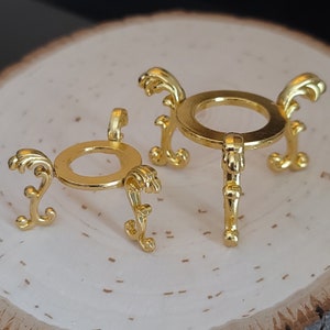 Può includere: Due supporti per sfere in metallo color oro con gambe curve e ornate e una parte superiore circolare. I supporti sono progettati per contenere sfere e hanno un'estetica decorativa e vintage. I supporti sono esposti su una superficie chiara e strutturata.