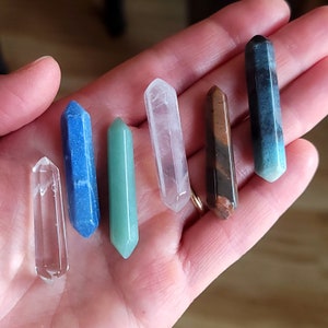 May include: A collection of six polished crystal wands in various colors, including clear, blue, green, pink, brown, and blue-green. Each wand is a slender, elongated shape with a pointed tip. The crystals are held in a hand.