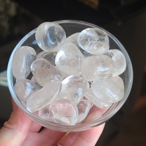 Op de afbeelding: Een heldere glazen kom gevuld met getrommelde heldere kwartsstenen. De stenen zijn glad, afgerond en doorschijnend, met een licht melkachtig uiterlijk. De kom wordt in een hand gehouden, met een donkere achtergrond.