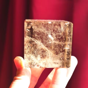May include: A translucent, cube-shaped crystal with a light golden-brown hue. The stone features visible internal fractures, natural inclusions, and a polished surface that catches the light, revealing a complex, textured pattern within the mineral.
