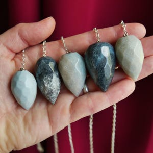 May include: Five faceted, teardrop-shaped stone pendants in varying shades of gray, blue-gray, and pale green. Each stone is attached to a silver-toned metal chain. The stones exhibit natural textures and polished surfaces with geometric facets.