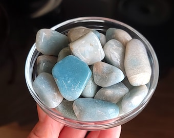 May include: A clear glass bowl filled with polished aquamarine stones. The stones vary in shades of blue and white, with some displaying a vibrant turquoise hue. The bowl is held in a hand, with a dark background.