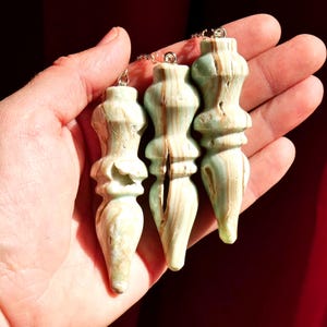 May include: Three carved stone pendants with a pale green and tan banded pattern. Each pendant features a decorative, multi-tiered spindle shape with a metal loop at the top for attachment. The stone displays natural variations and textures.
