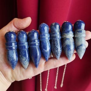May include: Seven blue sodalite crystal pendulums with a pointed, faceted shape and a rounded top section. Each stone features natural white marbling and is attached to a thin silver-colored metal chain, displaying a smooth, polished finish.