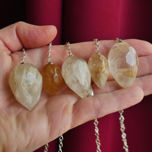 May include: Five faceted, teardrop-shaped crystal pendants in varying shades of translucent yellow, gold, and clear white. Each gemstone is attached to a thin, silver-colored metal chain, displaying natural internal inclusions and geometric cuts.
