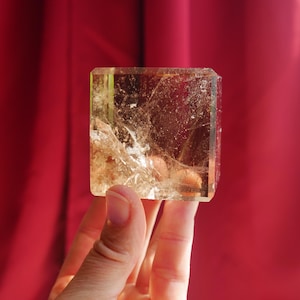 May include: A square-cut, translucent golden-brown crystal block featuring internal fractures, natural inclusions, and a polished surface. The mineral displays varying shades of amber and honey tones throughout its structure, catching light to reveal a complex, textured interior pattern.
