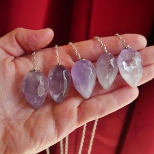 May include: Five faceted amethyst pendants, each with a teardrop shape and varying shades of purple, are suspended from silver-toned metal chains. The stones exhibit natural inclusions and translucent qualities, arranged in a row.